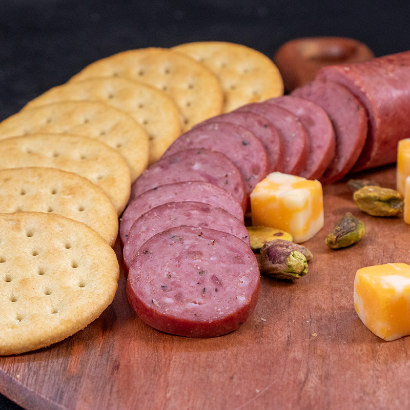 Slices of halal hot and spicy beef sausage is cut up and fanned out in a curve to the right. Large crackers are lined up to the left. A red dried pepper is to the right and some almonds are scattered around.