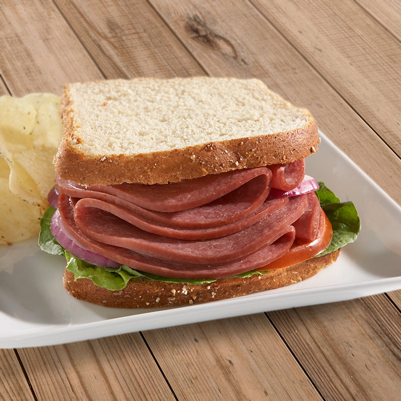 A Halal Beef salami sandwich with white bread red onion, tomato, and lettuce on a white plate with some potato chips behind to the left. 