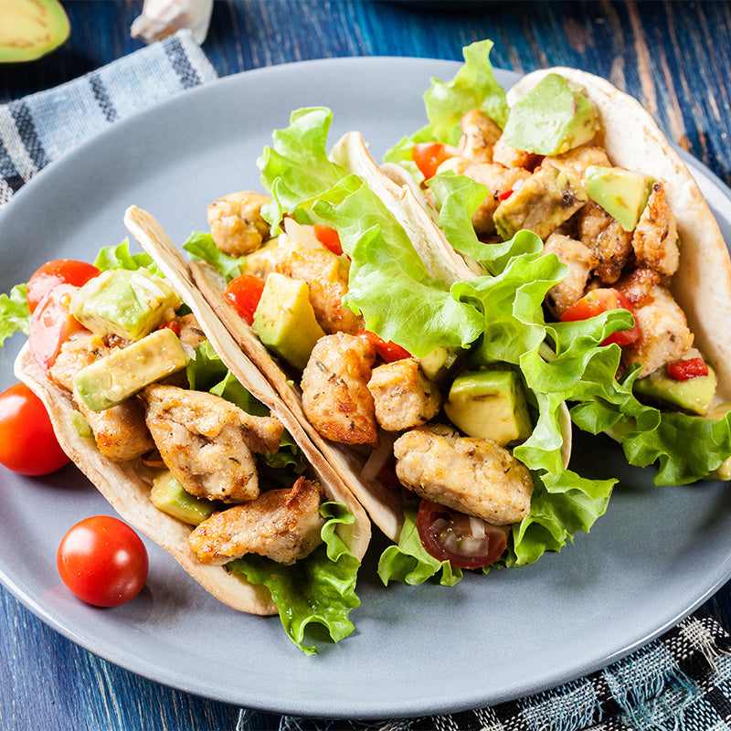 three tacos are lined up on a plate. In the tacos are diced chicken, avocado, tomatos and lettuce. 