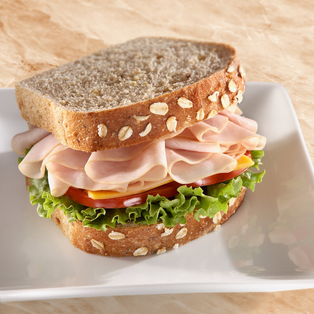 a turkey sandwich with oats baked into the top of the bread. The sandwich is piled high with Midamar halal turkey which is resting on a slice of cheese, tomatoes, and lettuce.