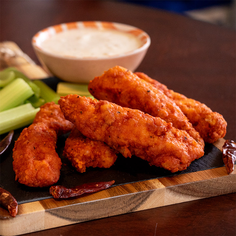 Midamar Spicy Chicken tenders are sitting on a cutting board with celery sticks, dried peppers, and a bowl of blue cheese.