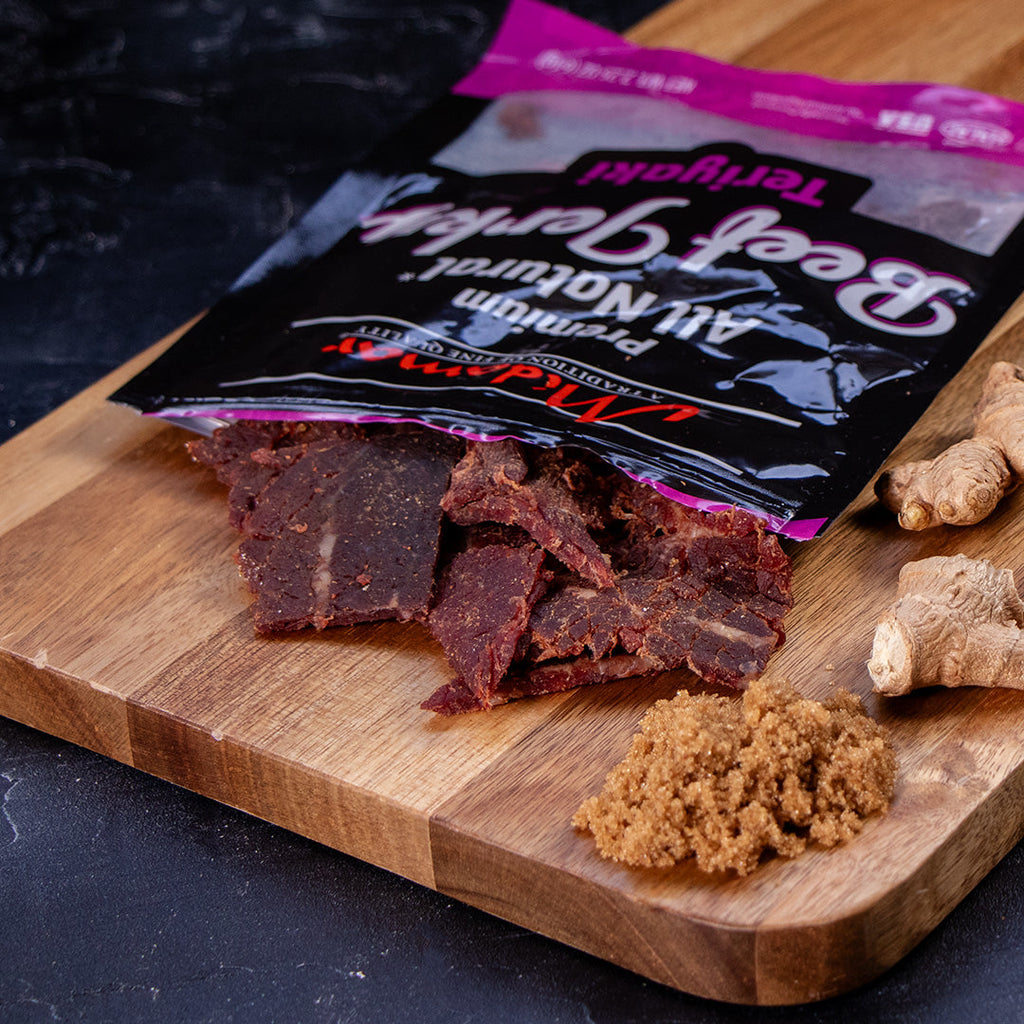 Midamar Halal Beef jerky Teriyaki pieces spill out of the bag onto a cutting board, a pile of brown sugar and ginger sit to the right of it.