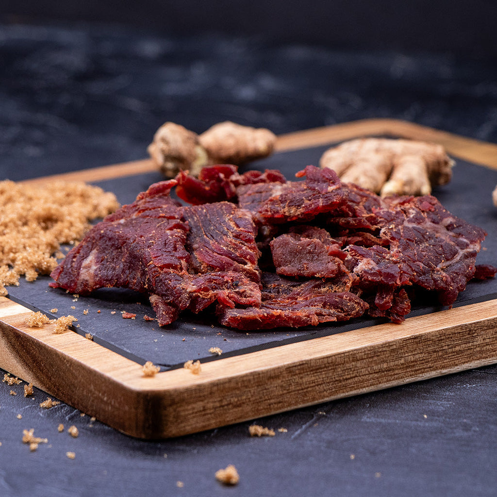 Midamar Halal Beef jerky Teriyaki pieces sit in a pile on a cutting board, brown sugar is sprinkled around and ginger is visible in the background.